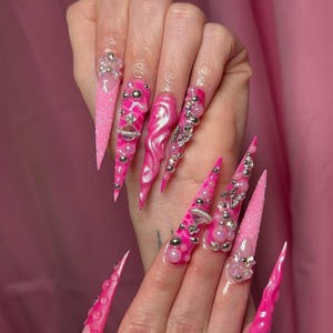 May include: Long, pointed, pink acrylic nails with various embellishments. Some nails have glitter, others feature silver beads, pearls, and decorative charms. The nails are a vibrant shade of pink.