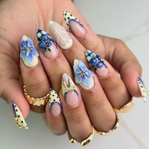 May include: A hand with long, almond-shaped nails featuring intricate nail art. Designs include 3D blue flowers, blueberries, lemon slices, polka dots, and checkered patterns. The nails are painted in shades of yellow, blue, and white, with gold accents.
