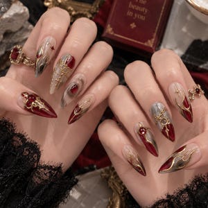 Puede incluir: Uñas postizas en forma de estilete con un diseño complejo de patrones marmolados en rojo oscuro y gris, detalles de cadenas de oro metálico y pequeñas gemas rojas incrustadas, adornadas con filigrana dorada y motivos en forma de estrella.