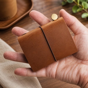 Op de afbeelding: Een klein, bruin leren notitieboekje in een hand. Het notitieboekje is rechthoekig met een messing knoopsluiting en een zwarte elastische band. Het leer heeft een getextureerde uitstraling. De achtergrond omvat een houten dienblad en groen.