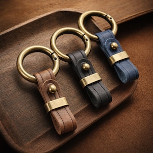 May include: Three leather keychains in brown, black, and blue, each with a brass ring and accents. The keychains are displayed on a wooden tray, showcasing the different colors and textures of the leather and metal.