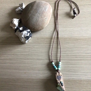 May include: A wooden pendant necklace with a brown cord. The pendant has a heart-shaped inlay and is decorated with turquoise and green beads. The necklace is displayed with natural stones on a wooden surface.