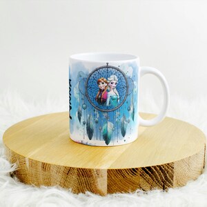 Op de afbeelding: Witte keramische mok met een dromenvanger ontwerp en twee cartoonfiguren. De mok heeft een blauwe aquarel achtergrond met de naam "Anna" verticaal afgedrukt. De mok staat op een houten ronde standaard.