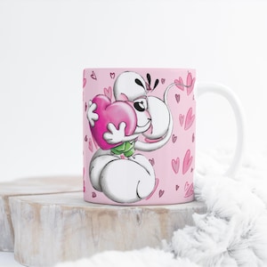 Könnte beinhalten: Eine weiße Keramik-Tasse mit einer Cartoon-Figur, die ein rosa Herz umarmt. Die Tasse hat einen rosa Hintergrund mit kleinen Herz-Illustrationen. Die Figur ist weiß mit schwarzen Akzenten und grünen Details. Die Tasse steht auf einer Holzoberfläche.