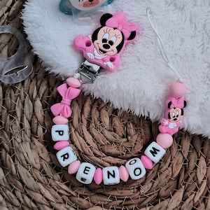Op de afbeelding: Een roze Minnie Mouse fopspeenclip met de naam "PENELOPE" gespeld in witte letterkralen en roze kralen. De clip heeft een Minnie Mouse figuur en een metalen clip. Een blauwe fopspeen met een berenontwerp is ook zichtbaar.