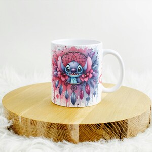 K&ouml;nnte beinhalten: Wei&szlig;e Keramiktasse mit einem farbenfrohen Aquarelldesign eines Zeichentrickcharakters und einem Traumf&auml;nger. Das Design umfasst rosa, blaue und violette Akzente. Die Tasse steht auf einem runden Holzst&auml;nder.