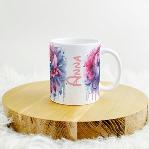 K&ouml;nnte beinhalten: Wei&szlig;e Keramik-Tasse mit einem Aquarelldesign mit einem Traumf&auml;nger und floralen Elementen in Rosa-, Blau- und Lilat&ouml;nen. Der Name "Anna" ist vertikal in Rosa geschrieben. Die Tasse steht auf einer runden Holzoberfl&auml;che.
