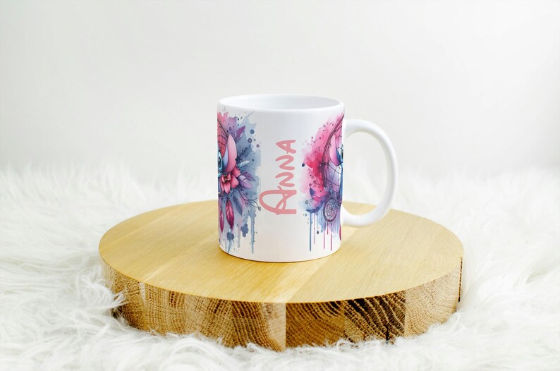 K&ouml;nnte beinhalten: Wei&szlig;e Keramik-Tasse mit einem Aquarelldesign mit einem Traumf&auml;nger und floralen Elementen in Rosa-, Blau- und Lilat&ouml;nen. Der Name "Anna" ist vertikal in Rosa geschrieben. Die Tasse steht auf einer runden Holzoberfl&auml;che.