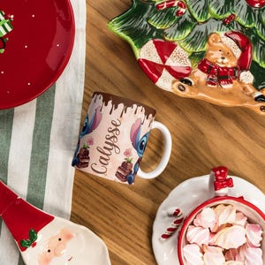 May include: A festive holiday scene with a red plate featuring gingerbread men and a gift, a Christmas tree-shaped serving dish, and a mug with a cartoon character design. A Santa-shaped dish and a bowl of marshmallows complete the arrangement.