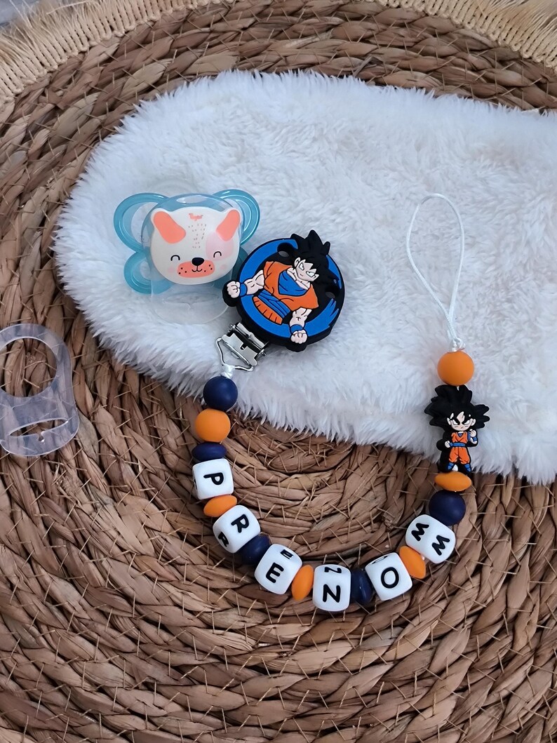 May include: A baby pacifier and a pacifier clip with the name "PREZMO" spelled out in white lettered beads. The pacifier has a blue and white design with a bear face. The clip features a Dragon Ball Z character.