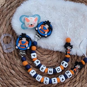 May include: Baby pacifier set featuring a blue and white bear-shaped pacifier and two pacifier clips. The clips have a blue and orange bead design with the name "JULIA PRENZO" spelled out in white letter beads. Each clip has a Goku character design.