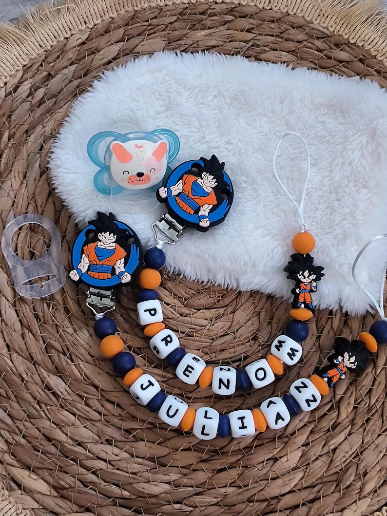 May include: Baby pacifier set featuring a blue and white bear-shaped pacifier and two pacifier clips. The clips have a blue and orange bead design with the name "JULIA PRENZO" spelled out in white letter beads. Each clip has a Goku character design.