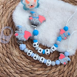 Peut inclure: Une sucette pour bébé et deux attaches-sucettes sur le thème de Dumbo l'éléphant. La sucette est bleu clair avec un visage d'ours. Les clips et la chaîne épellent "BABY" avec des perles de lettres blanches.