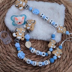 Könnte beinhalten: Baby-Schnuller und Schnullerclips in einer neutralen Farbpalette. Der Schnuller hat ein blau-weißes Design mit einem Fuchskopf. Die Clips haben löwenförmige Silikonperlen, Holzperlen und die Namen "NINO" und "TOM".
