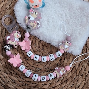 Puede incluir: Un conjunto de accesorios para bebés, que incluye un chupete azul y blanco, dos clips para chupete y una almohadilla de felpa blanca. Los clips para chupete presentan cuentas rosas, detalles de mariposas y el nombre "CHLOÉ" deletreado con cuentas de letras blancas.