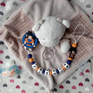 May include: A baby comfort blanket with a plush sheep head, a pacifier, and a pacifier clip featuring a Dragon Ball Z character. The clip has the name "PREZOM" spelled out in letter beads. The blanket is beige with a heart pattern.