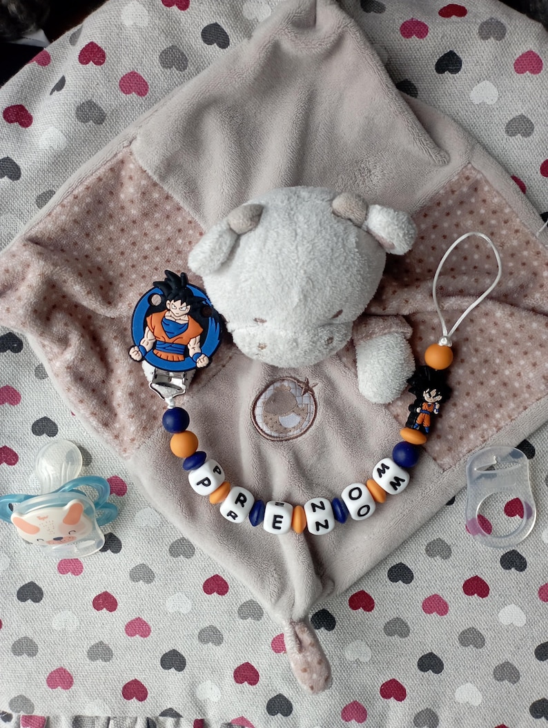 May include: A baby comfort blanket with a plush sheep head, a pacifier, and a pacifier clip featuring a Dragon Ball Z character. The clip has the name "PREZOM" spelled out in letter beads. The blanket is beige with a heart pattern.