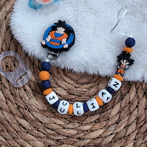 May include: A baby pacifier clip with the name "JULIAN" spelled out in white letter beads. The clip features a cartoon image of a character in an orange and blue outfit. The clip is attached to a blue and orange beaded chain, and a blue pacifier with a bear face.