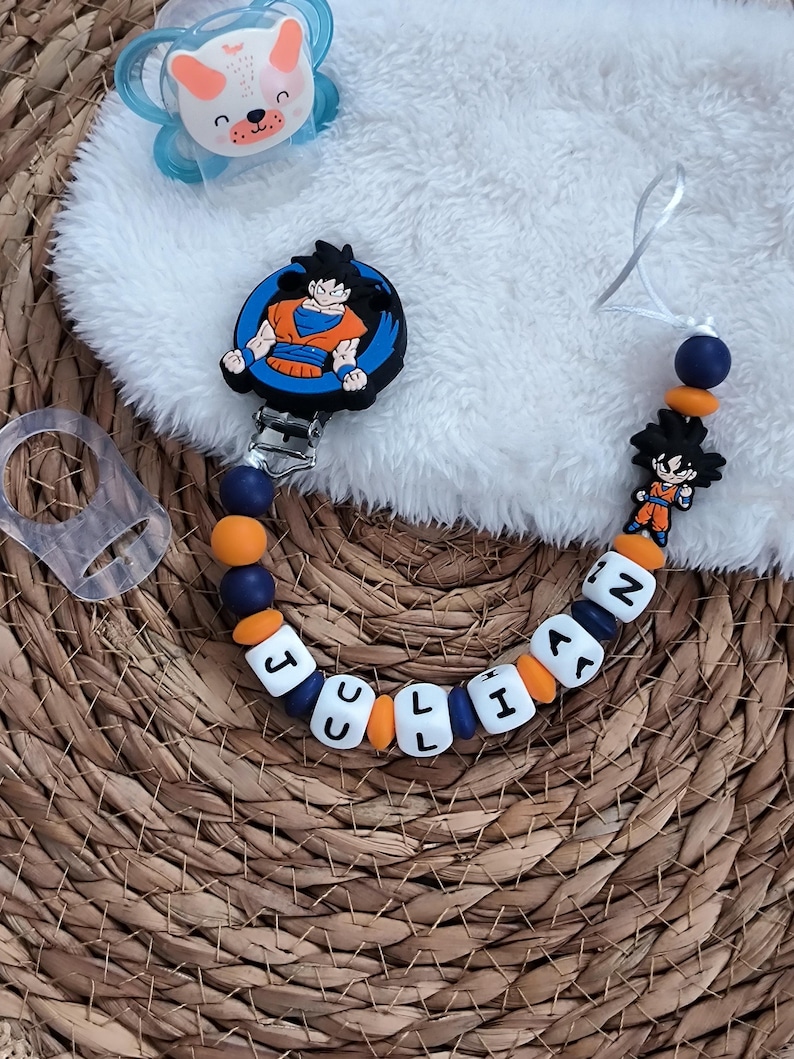 May include: A baby pacifier clip with the name "JULIAN" spelled out in white letter beads. The clip features a cartoon image of a character in an orange and blue outfit. The clip is attached to a blue and orange beaded chain, and a blue pacifier with a bear face.