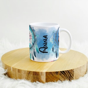 Op de afbeelding: Witte keramische mok met een dromenvanger ontwerp met blauwe en turquoise aquarel accenten. De naam "Anna" staat verticaal in zwart schrift. De mok staat op een ronde houten ondergrond.