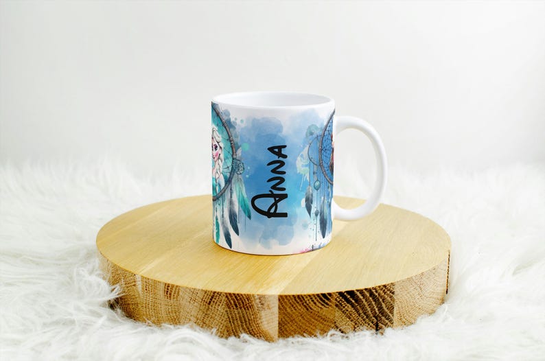 Op de afbeelding: Witte keramische mok met een dromenvanger ontwerp met blauwe en turquoise aquarel accenten. De naam "Anna" staat verticaal in zwart schrift. De mok staat op een ronde houten ondergrond.