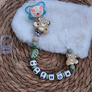 Könnte beinhalten: Ein Baby-Schnuller- und Schnullerclip-Set. Der Schnuller ist blau mit einem weißen und orangefarbenen Hasenmuster. Der Clip hat ein Löwen-Design, grüne Perlen und den Namen "RENOM" in weißen Buchstaben auf schwarzen Perlen.