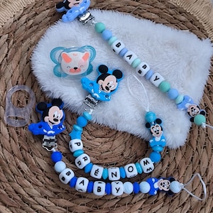 Op de afbeelding: Een verzameling babyaccessoires met een Mickey Mouse-thema. De set bevat blauwe fopspeenclips met het woord "BABY" in witte letters, een blauwe fopspeen en een witte, zachte deken.