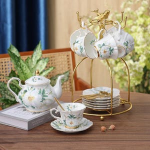 Può includere: Un servizio da tè in porcellana bianca con motivi floreali. Il set comprende una teiera, tazze, piattini e un supporto in metallo dorato per le tazze. Le tazze e la teiera sono decorate con fiori bianchi e foglie verdi.