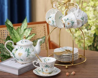May include: A white porcelain tea set with floral designs. The set includes a teapot, teacups, saucers, and a gold-colored metal stand for the cups. The teacups and teapot are decorated with white flowers and green leaves. A small gold spoon rests in one of the teacups.