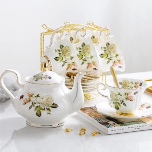 Puede incluir: Un juego de té de porcelana blanca con un diseño floral de rosas. El juego incluye una tetera, tazas, platillos y un soporte de metal dorado. Las tazas están colgadas en el soporte. Una cuchara dorada descansa en una de las tazas.