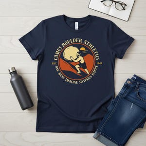 May include: Navy blue t-shirt with a circular graphic featuring a figure pushing a large ball up a hill, with the text "Camus Boulder Athletics" and "One Must Imagine Sisyphus Happy". A black water bottle and jeans are also visible.