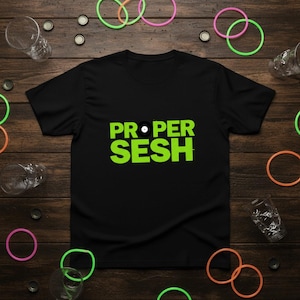 May include: Black t-shirt with the words "PROPER SESH" in bright green, with a vinyl record graphic. The shirt is surrounded by colorful rubber bands and empty glasses on a wooden surface.