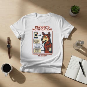 Puede incluir: Camiseta blanca con un gráfico de un perro y el texto "PAVLOV'S NOTIFICATION" y "CLASSICALLY CONDITIONED TO ANXIETY". El diseño incluye un icono de teléfono, flechas y cuadros de texto. El perro es marrón y rojo.