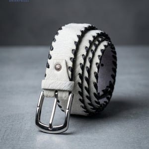 May include: A white hair-on-hide belt featuring black whipstitch detailing along the edges. The belt includes a silver-toned metal buckle and a single matching white hair-on-hide keeper loop. The texture of the material is visibly coarse.
