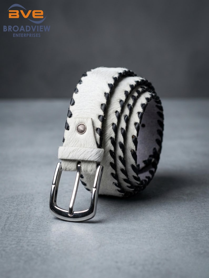May include: A white hair-on-hide belt featuring black whipstitch detailing along the edges. The belt includes a silver-toned metal buckle and a single matching white hair-on-hide keeper loop. The texture of the material is visibly coarse.