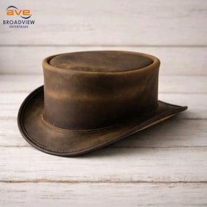 Könnte beinhalten: Ein brauner Lederzylinder mit breiter Krempe. Der Hut hat eine zylindrische Krone und eine leicht gebogene Krempe. Der Hut ist aus einem Leder mit Used-Look gefertigt. Der Hut ist ein klassischer Stil.