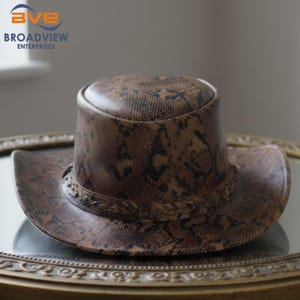 Puede incluir: Un sombrero con estampado de piel de serpiente en marrón y negro, con una parte superior plana y un ala ancha y curva. El sombrero presenta una banda trenzada decorativa que rodea la base de la copa, mostrando una textura reptiliana.
