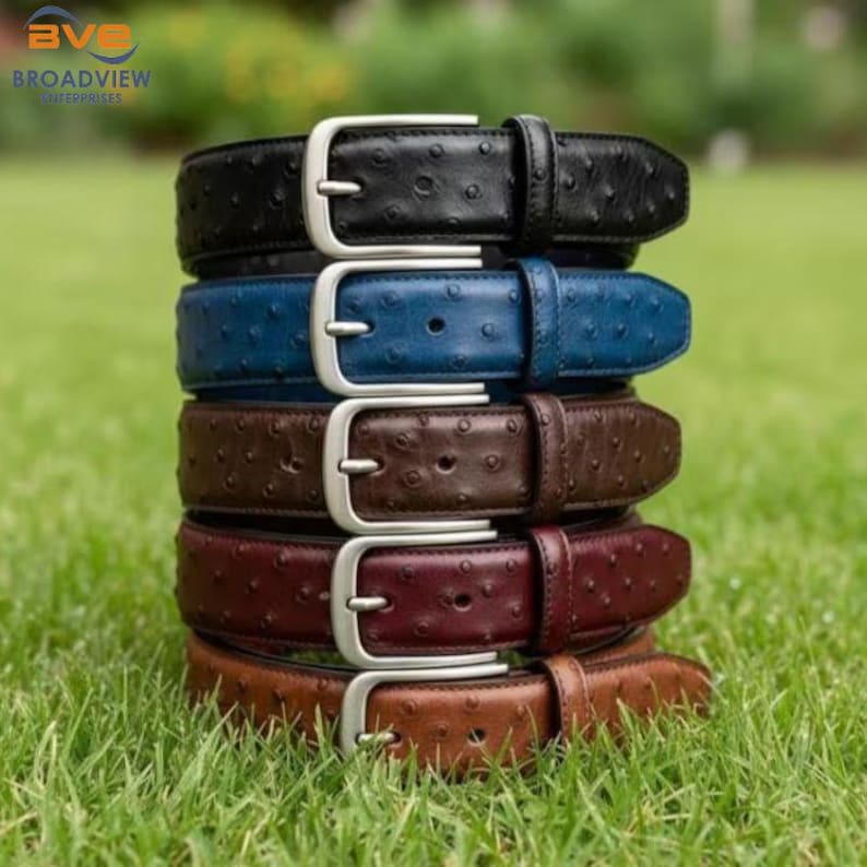 May include: A stack of five leather belts in various colors: black, blue, brown, burgundy, and tan. Each belt has a silver-colored buckle and a textured surface. The belts are arranged on a bed of green grass.