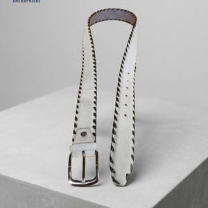 May include: A white belt with a textured hair-on-hide finish and dark brown stitched edges. The belt features a silver-toned metal buckle and a series of adjustment holes along the strap, showcasing a distinct contrast between the white surface and dark trim.