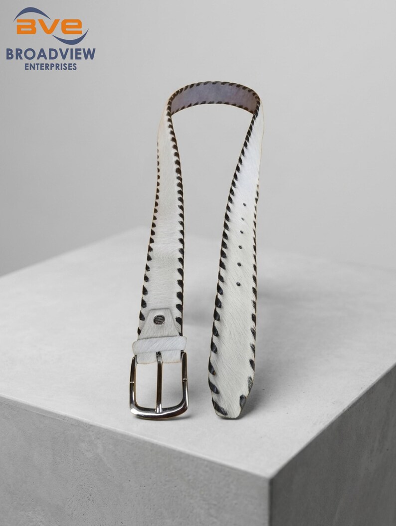 May include: A white belt with a textured hair-on-hide finish and dark brown stitched edges. The belt features a silver-toned metal buckle and a series of adjustment holes along the strap, showcasing a distinct contrast between the white surface and dark trim.