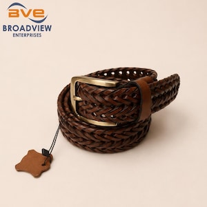 Op de afbeelding: Een donkerbruine gevlochten riem met een rechthoekige metalen gesp in antieke messingkleur. De riem is voorzien van een gladde bruine leren lus en een klein, onregelmatig gevormd bruin leren label dat vastzit met een dun zwart koordje.