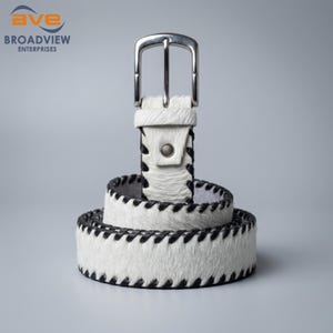 May include: A white hair-on-hide belt featuring black stitched edges and a silver-toned metal buckle. The belt is coiled in a circular shape, displaying a textured surface with a distinct pattern of dark thread along the top and bottom borders.