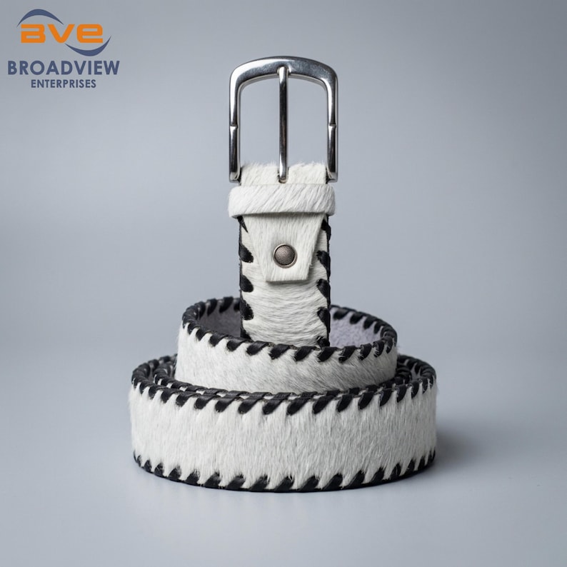 May include: A white hair-on-hide belt featuring black stitched edges and a silver-toned metal buckle. The belt is coiled in a circular shape, displaying a textured surface with a distinct pattern of dark thread along the top and bottom borders.