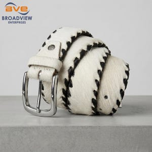 May include: A coiled belt made of white hair-on hide with black spotted patterns and black whipstitch detailing along the edges. The belt features a polished silver-tone metal buckle and a matching leather keeper loop for a secure closure.