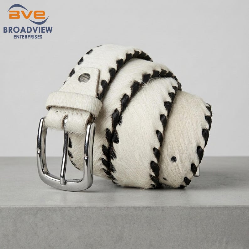 May include: A coiled belt made of white hair-on hide with black spotted patterns and black whipstitch detailing along the edges. The belt features a polished silver-tone metal buckle and a matching leather keeper loop for a secure closure.