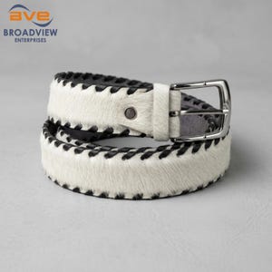 May include: A white hair-on-hide belt featuring black whipstitch detailing along the edges. The belt includes a silver-toned metal buckle and a single matching white hair-on-hide belt loop, with a dark interior lining visible on the strap.
