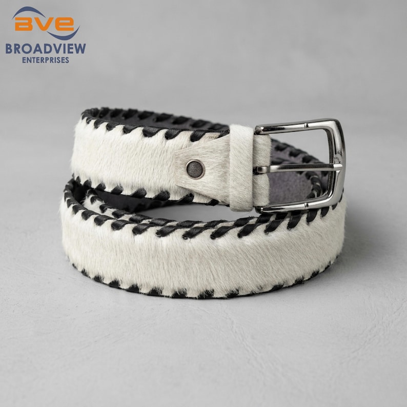 May include: A white hair-on-hide belt featuring black whipstitch detailing along the edges. The belt includes a silver-toned metal buckle and a single matching white hair-on-hide belt loop, with a dark interior lining visible on the strap.