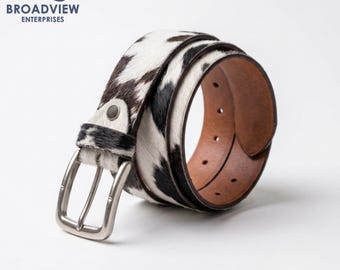 May include: A belt featuring a cowhide pattern with black and white fur on the exterior and a smooth brown leather interior. The belt includes a silver-toned metal buckle, a matching leather loop, and a series of adjustment holes along the strap.