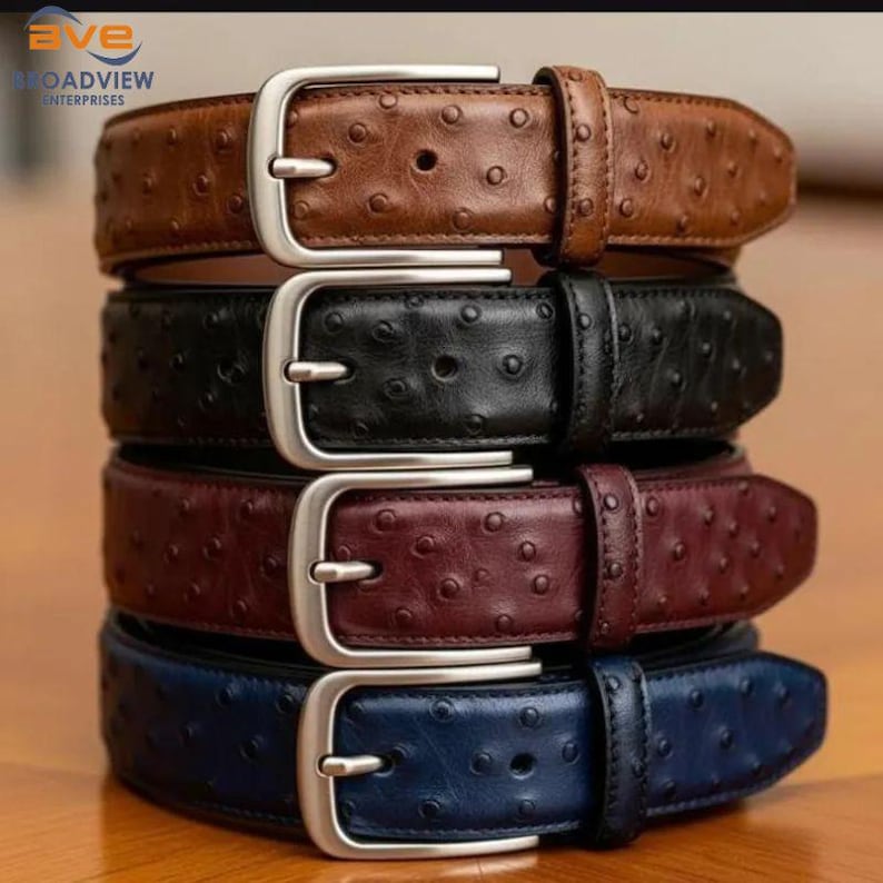 May include: A stack of four leather belts in various colors: brown, black, burgundy, and navy blue. Each belt features a silver-colored buckle and a textured surface with small, evenly spaced dimples. The belts are stacked on top of each other.