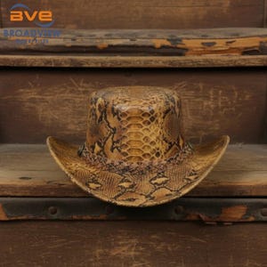 Puede incluir: Un sombrero de vaquero con un patrón de piel de serpiente en tonos bronceado y marrón en toda la copa y el ala. El sombrero incluye una banda trenzada alrededor de la base de la copa y dos pequeños detalles metálicos en los lados del ala.